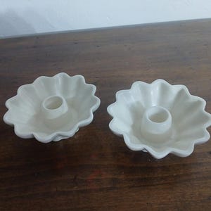 May include: Two off-white ceramic candle holders. Each holder has a flower-shaped design with a central cylindrical cup for holding a candle. The holders are placed on a dark wooden surface.