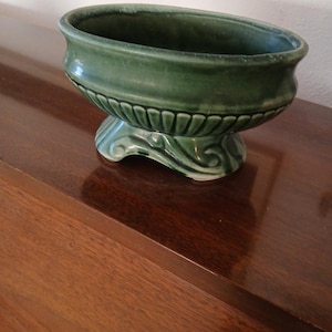 May include: A green ceramic planter with an oval bowl shape and a decorative base. The planter has a glossy finish and is sitting on a wooden surface. The base features a scroll design.