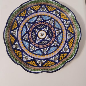 May include: A round ceramic plate with a scalloped edge featuring a complex hand-painted pattern. The design includes concentric circles of deep blue, yellow, and green, with intricate floral motifs, geometric shapes, and dotted textures.