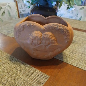 May include: Heart-shaped terracotta planter with embossed cherub and floral designs. The planter is a warm, earthy tone and sits on a wooden table. Decorative vase and greenery are in the background.