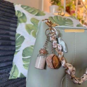 Peut inclure: Un sac à main vert clair avec des accessoires dorés et un porte-clés décoratif. Le porte-clés comprend une petite bouteille en verre avec un bouchon en liège, un charm en forme de baseball et une chaîne en ruban de satin. Un coussin avec un motif de feuilles vertes est en arrière-plan.