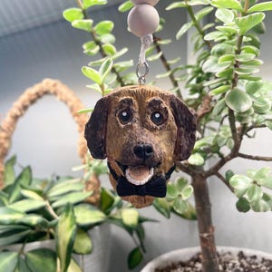 May include: A handcrafted dog head ornament with a black bow tie. The dog's head is brown with dark brown ears and a smiling expression. The ornament hangs from a string with wooden beads, perfect for a pet lover.