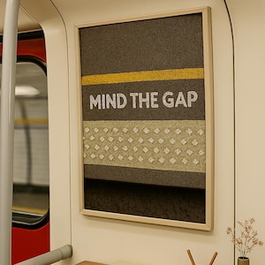 Puede incluir: Obra de arte enmarcada con el texto "MIND THE GAP" en letras blancas. La obra presenta un fondo marrón con una franja amarilla y una sección estampada. La escena está ambientada en un vagón de tren, con una mesa de madera y objetos decorativos.