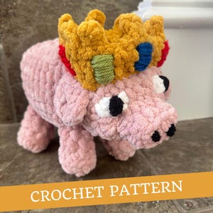 May include: A pink crocheted pig wearing a yellow, red, blue and green crocheted crown. The pig has black eyes and a black nose.