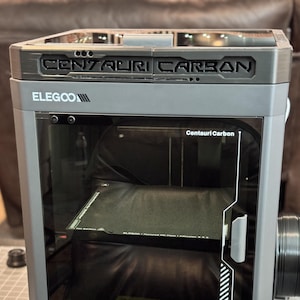 Puede incluir: Una impresora 3D gris ELEGOO Centauri Carbon con una puerta frontal transparente. La impresora tiene el texto "CENTAURI CARBON" en la parte superior e incluye un pequeño panel de control con una pantalla que muestra varias configuraciones. La impresora está sobre una mesa.