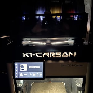 May include: A black and white 3D printer with the text "X1-CARBON" illuminated on the front. A small screen displays a user interface. The printer is in a dimly lit environment.