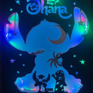 Op de afbeelding: Een zwart omrand lichtgevend bord met het personage Stitch uit Lilo & Stitch. Het bord heeft een blauw silhouet van Stitch met het woord "Ohana" erboven. Het bord wordt verlicht door kleurrijke LED-verlichting, sterren en een palmboom.