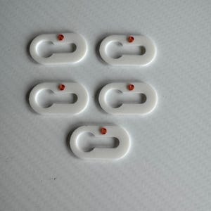 Op de afbeelding: Vijf witte, ovale plastic connectoren met een kleine rode kraal in het midden. Ze hebben een C-vormige opening aan één kant en een klein gaatje aan de andere kant.