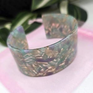 Op de afbeelding: Een brede, open manchetarmband met een gemarmerd effect in groen, paars en beige tinten. De armband ligt op een roze stoffen zakje, met groene bladeren op de achtergrond. De manchet is ongeveer 5 cm breed.