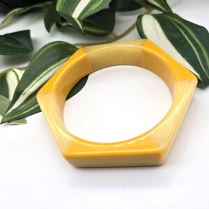 May include: A hexagonal bangle bracelet featuring alternating segments of opaque mustard yellow and translucent pale yellow resin. The geometric design has a smooth, polished finish with a consistent thickness throughout the entire circular band.