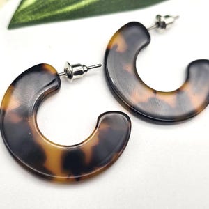 May include: A pair of C-shaped hoop earrings featuring a brown and black tortoiseshell pattern. The earrings are constructed with a smooth, polished finish and include silver-toned metal post backings for secure attachment to the earlobe.