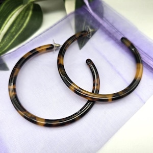 Puede incluir: Un par de pendientes de aro grandes con un patrón de carey. Los pendientes son marrones y negros, con un diseño circular. Están sobre una superficie blanca y morada, con hojas verdes y blancas en el fondo.