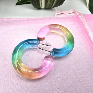 Könnte beinhalten: Ein Paar bunte Creolen. Die Ohrringe bestehen aus durchscheinendem Material mit einem Farbverlauf aus Rosa, Orange, Gelb, Grün und Blau. Die Ohrringe liegen auf einer rosa Oberfläche.