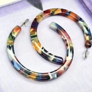 Puede incluir: Un par de pendientes de aro coloridos. Los pendientes tienen un patrón veteado con tonos de naranja, verde, azul y morado. Los pendientes están colocados sobre un fondo de tela blanca. Un accesorio de moda.