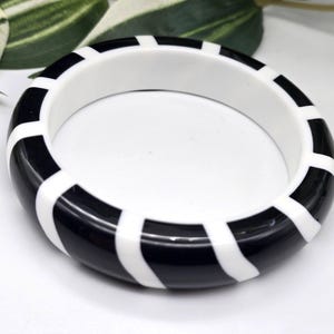 以下が含まれることがあります： 黒と白のストライプ柄のバングルブレスレット。太くて丸いブレスレットは、光沢のある黒と白の交互のバンドが特徴です。どんな服装にも合わせやすい、印象的なアクセサリーです。