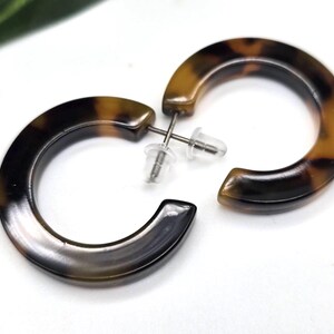 May include: A pair of tortoiseshell-patterned hoop earrings. The earrings have a C-shaped design with a post and backing. The color pattern is a mix of brown and black, with a glossy finish. The earrings are set against a white background.