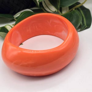 Op de afbeelding: Een dikke, gladde, feloranje armband met een afgeronde, organische vorm. Het accessoire heeft een glanzende, effen afwerking en een breed, hol midden, wat een eenvoudig en gedurfd ontwerp laat zien dat uniform is in zijn levendige kleur.