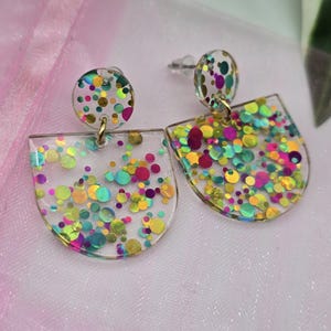 Puede incluir: Un par de pendientes de resina transparente con purpurina de colores. Cada pendiente tiene un pequeño perno redondo y un colgante semicircular más grande. La purpurina incluye tonos de rosa, amarillo, verde y azul, creando un diseño llamativo.