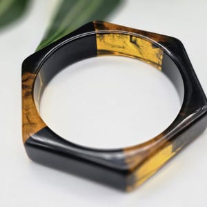 Könnte beinhalten: Sechseckiges Armreif-Armband in Schwarz und Bernstein. Das geometrische Armband zeichnet sich durch einen auffälligen Farbkontrast und eine einzigartige Form aus. Die Bernsteinfarbenen Abschnitte haben einen Marmoreffekt und verleihen diesem Accessoire zusätzliches Interesse.