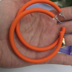 Può includere: Un paio di orecchini a cerchio arancioni brillanti. Gli orecchini sono di un colore vivace e uniforme e hanno un design liscio e arrotondato. I perni degli orecchini sono argentati. Un accessorio di moda audace.