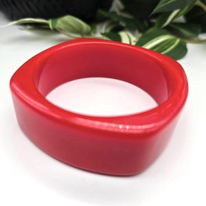 May include: A bright red, square-profile bangle. The bracelet has a smooth, glossy surface and a wide band. The bold colour and geometric design make it a striking accessory. The bracelet is a fashion item.