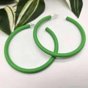 May include: A pair of bright green hoop earrings. The earrings are a solid, vibrant green color and have a smooth, matte finish. The earrings are round and have a simple, minimalist design. The earrings are shown against a white background with green and white leaves.