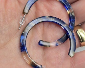 May include: A pair of hoop earrings with a translucent design. The earrings feature a mix of blue, brown, and clear colors. The earrings have a post-back closure. The earrings are held in a hand.