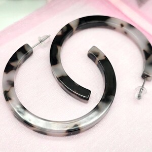 May include: A pair of hoop earrings with a marbled pattern in black, gray, and white. The earrings are made of a smooth material and have a post-back closure. The earrings are displayed on a pink surface.