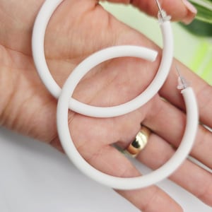 May include: A pair of large, white hoop earrings. The earrings are a solid, matte white colour and have a simple, minimalist design. The hoops are held in a hand, showcasing their size and shape. The earrings are a classic accessory.