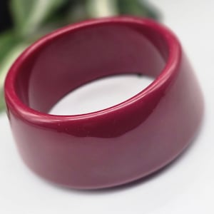May include: A wide, burgundy-coloured bangle bracelet. The smooth, glossy surface reflects light, highlighting its rounded shape. This bold accessory is a statement piece, perfect for adding a pop of colour to any outfit.