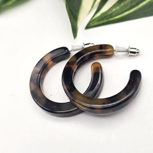 May include: A pair of hoop earrings featuring a brown and black tortoiseshell pattern. The earrings are circular in shape with a smooth, polished finish and include silver-toned metal posts with clear plastic earring backs for secure closure.