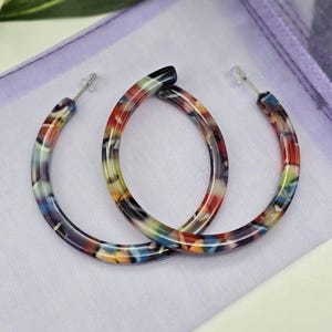 May include: A pair of colourful hoop earrings. The earrings have a marbled pattern with shades of red, blue, yellow, and purple. The earrings are made of a lightweight material and have a silver-coloured post. The earrings are displayed on a white surface.