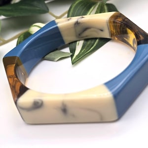 Op de afbeelding: Een geometrische armband met een zeshoekige vorm. De armband heeft secties van blauwe, crème en bruine hars. Een modeaccessoire.