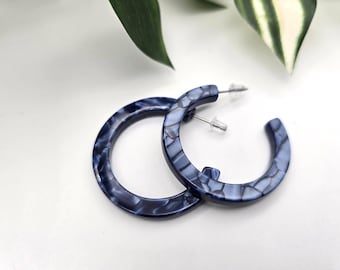 May include: A pair of blue and white marbled hoop earrings. The earrings are made of a thick, rounded material and have a post closure. The earrings are set against a white background with green leaves.