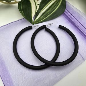 Puede incluir: Un par de pendientes de aro negros grandes. Los pendientes son circulares y están hechos de un material negro mate. Se exhiben sobre una tela lavanda con hojas verdes y blancas en el fondo.