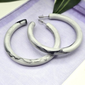 May include: A pair of white hoop earrings with a marbled black and gray design. The earrings are circular and have a post closure. They are displayed on a lavender fabric.