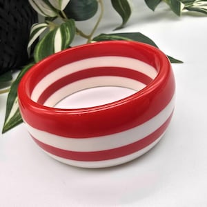 May include: A red and white striped bangle bracelet. The wide, oval-shaped bracelet features alternating bands of glossy red and white. The bracelet is a bold, eye-catching accessory.