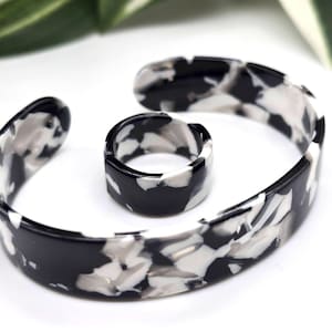 May include: A black and white marbled cuff bracelet and matching ring. The jewelry has a bold, geometric pattern with a mix of black, white, and gray tones. The bracelet is open-ended, and the ring is a thick band.