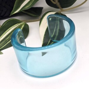 Könnte beinhalten: Ein breites, durchscheinendes hellblaues Armband aus glattem, poliertem Acrylmaterial. Das Armband hat ein offenes C-förmiges Design mit abgerundeten Kanten und zeigt ein klares, glasartiges Aussehen, das Licht durch das Material hindurchscheinen lässt.