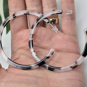 May include: A pair of large hoop earrings with a marbled pattern in black and white. The earrings are held in a hand, showcasing their size and design. The earrings have a post and backing for secure wear.