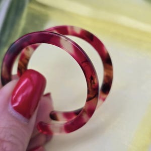 Op de afbeelding: Twee ronde oorringen in schildpadstijl. De oorbellen hebben een mix van rode, bruine en crèmekleurige kleuren. De oorbellen worden tegen een gele achtergrond gehouden.