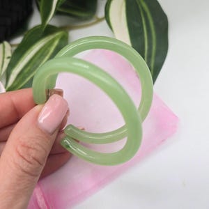 Op de afbeelding: Een paar lichtgroene oorringen. De oorbellen hebben een dubbel-hoepel ontwerp, met een gladde, glanzende afwerking. De oorbellen worden tegen een roze doek gehouden, met groen-witte bladeren op de achtergrond. Sieraden.