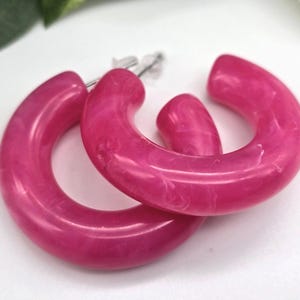May include: A pair of bright pink hoop earrings. The earrings are made of a glossy, marbled material. The earrings are a bold, eye-catching accessory.