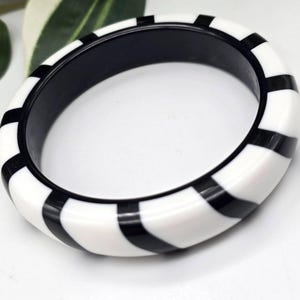 May include: A white and black striped bangle bracelet. The bracelet has a rounded square shape with alternating black and white stripes. The interior of the bracelet is black. The bracelet is a fashion accessory.