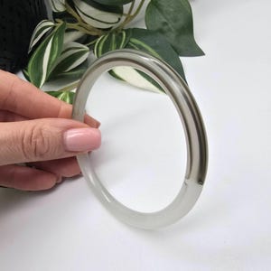 May include: A clear, round bangle bracelet with a grey stripe. The bracelet is held by a hand with pink nail polish. The background includes green and white leaves.