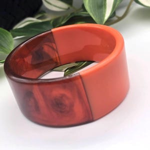 Könnte beinhalten: Ein breites, klobiges Armband in Rot- und Orangetönen. Das Armband ist in zwei Abschnitte unterteilt, einer mit marmoriertem, holzähnlichem Aussehen und der andere einfarbig, glatt orange. Ein auffälliges Accessoire.