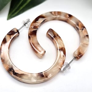 May include: A pair of hoop earrings with a marbled pattern in shades of brown, tan, and cream. The earrings have a post-back closure and are set against a white background. The earrings are a fashion accessory.