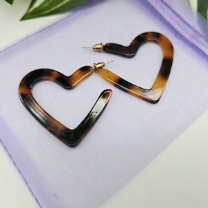 May include: A pair of heart-shaped earrings in a tortoiseshell pattern. The earrings are made of dark brown and tan material and have gold-coloured posts. They are displayed on a light purple fabric.