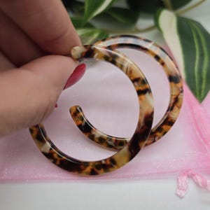 May include: A pair of large, hoop earrings with a tortoiseshell pattern. The earrings are a mix of brown, tan, and cream colors. They are held against a pink mesh bag and greenery.