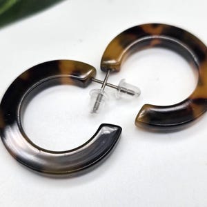 Könnte beinhalten: Ein Paar Creolen mit Schildpattmuster. Die Ohrringe haben eine halbkreisförmige Form mit Stecker und Verschluss. Das Farbmuster ist eine Mischung aus Braun und Beige.